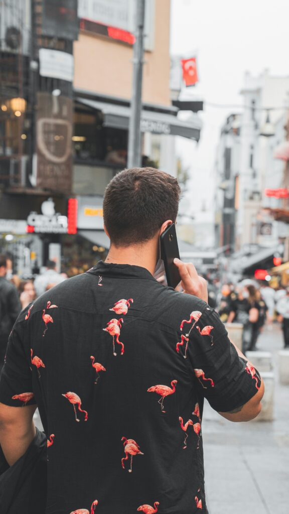 Homem falando ao celular em rua movimentada, usando camisa preta com estampa de flamingos.