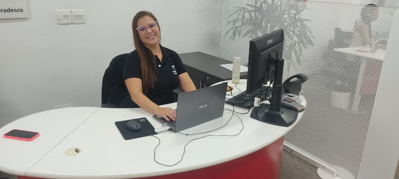 “Mulher sorridente, usando óculos e camisa preta com logotipo, sentada em uma mesa de escritório com notebook e monitor.”